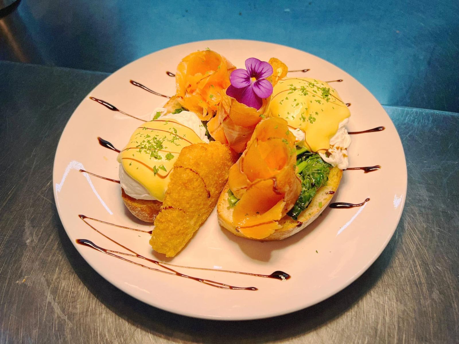 Huevos pochados con salsa holandesa, salmón ahumado y espinaca sobre pan tostado, con papa rösti y una flor comestible